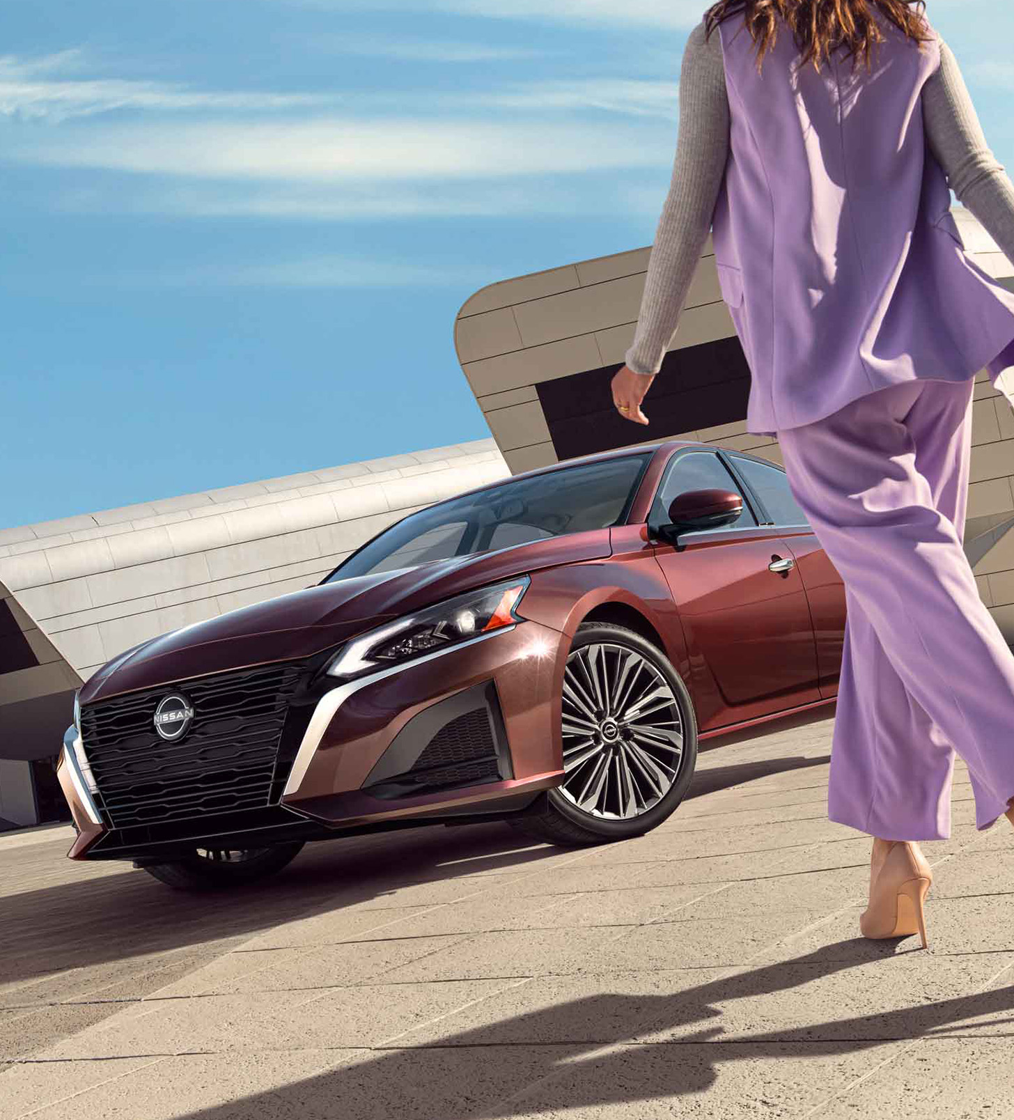 Una mujer camina hacia el Nissan Altima 2024, cuyo diseño brillante en color Rojo Burdeos se destaca contra una arquitectura moderna.