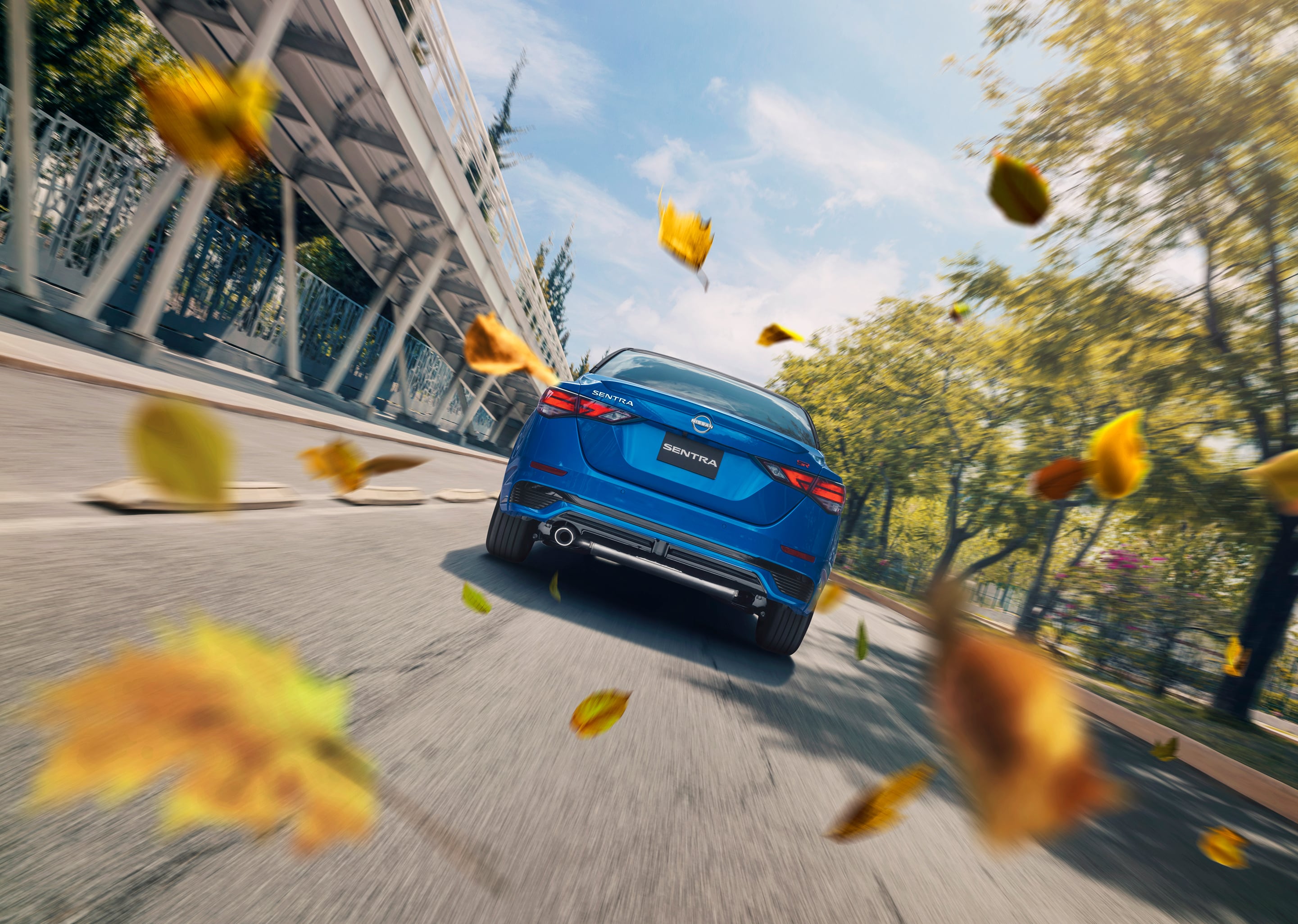 Vista trasera del Nissan Sentra 2025 color azul recorriendo un entorno urbano con hojas de otoño al viento, resaltando su seguridad y desempeño en carretera.