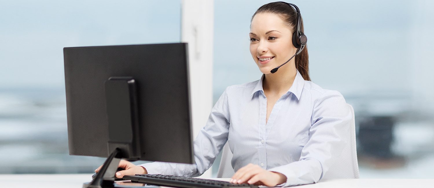 Una operadora de call center sonríe amablemente mientras mira su monitor, con una mano teclea y con la otra sostiene el mouse de su computadora, el fondo está fuera de foco​