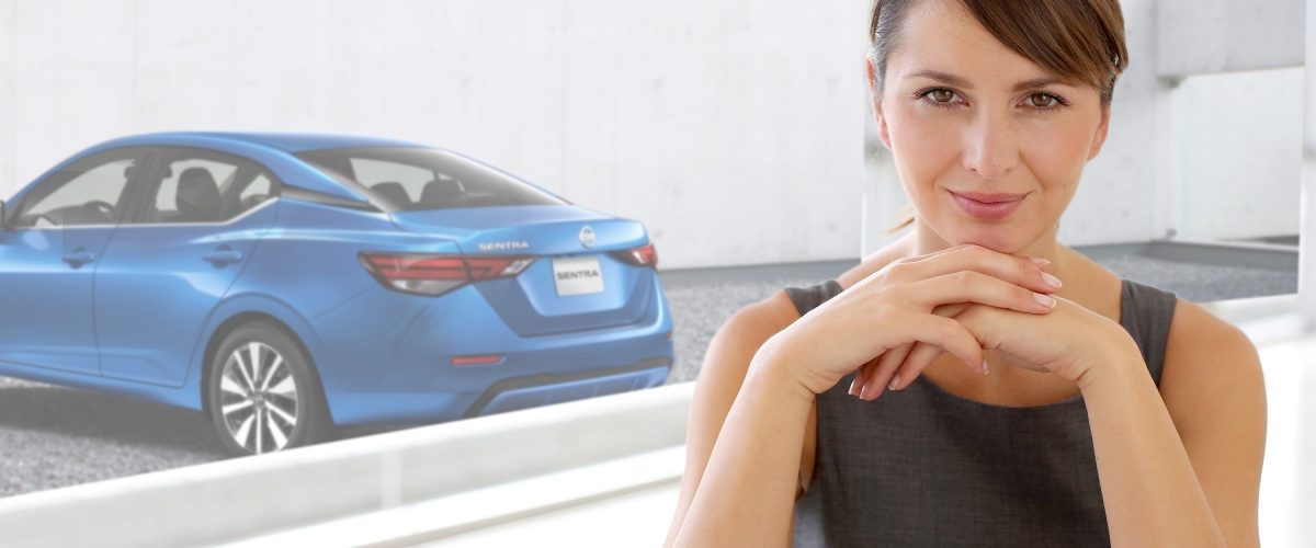 Una mujer mira decidida directamente a cámara con un Nissan Sentra estacionado en la parte exterior del lugar​